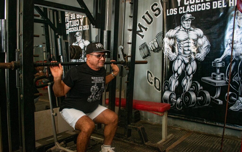 Entrenador veterano ejecutando sentadilla en máquina Smith en el gimnasio del barrio Los Cerezos. 