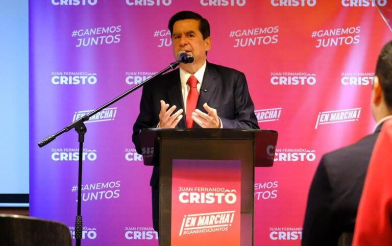 Juan Fernando Cristo interviene en tarima durante un evento público, frente a un fondo con su eslogan de campaña.